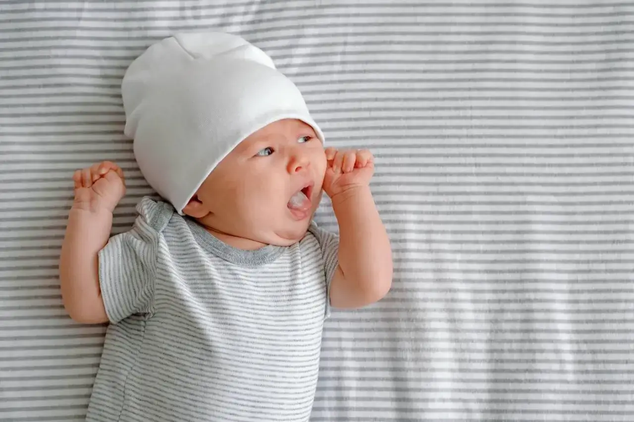 Baby mit wei&szlig;er M&uuml;tze und gestreiftem Strampler zeigt den Suchreflex, die Augen sind weit ge&ouml;ffnet.