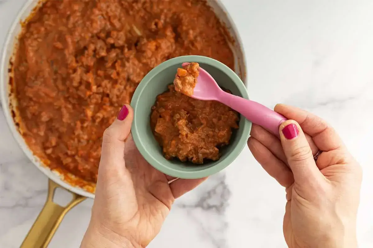 Eine Hand f&uuml;llt eine Sch&uuml;ssel mit Linsenbolognese f&uuml;r ein Baby. Eine rosa L&ouml;ffel liegt bereit.