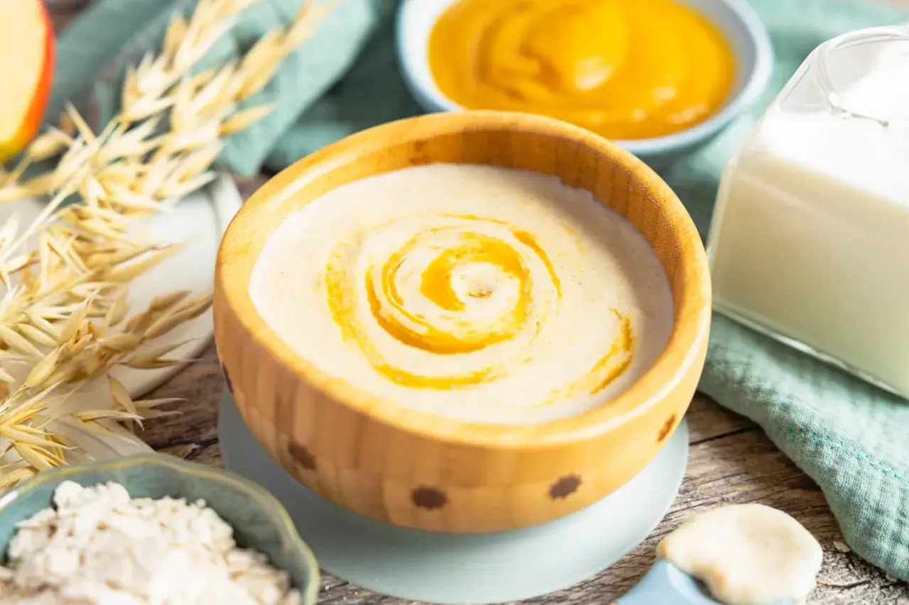 Ein cremiger Milch-Getreide-Brei im Holzsch&auml;lchen, garniert mit &Ouml;l. Zutaten wie Haferflocken und ein Glas Milch deuten auf ein einfaches Rezept hin.