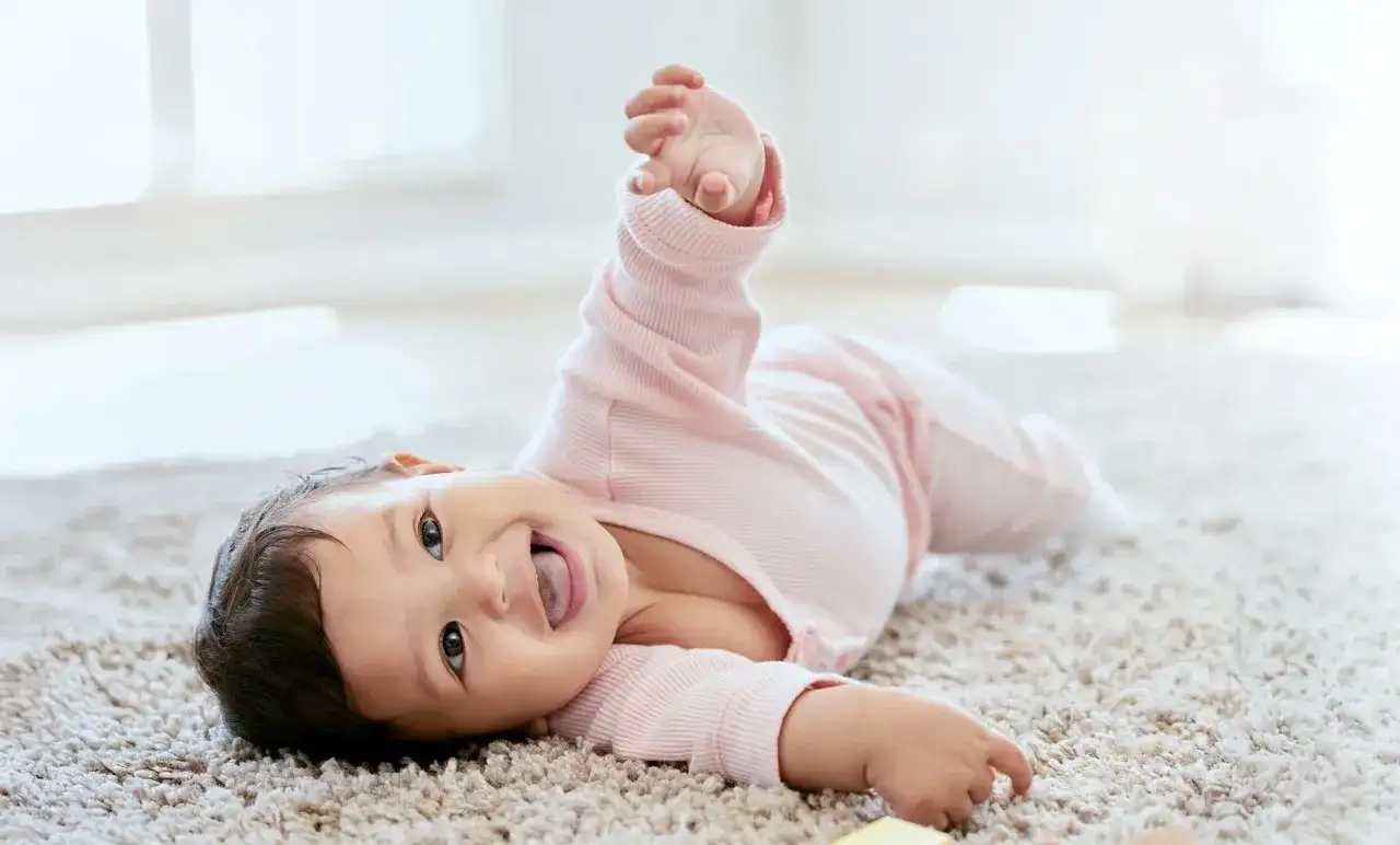 Ein Baby in rosa Strampler liegt auf einem Teppich und versucht, sich zu drehen. Ab wann drehen sich Babys? Dieses kleine Wunder übt schon fleißig!
