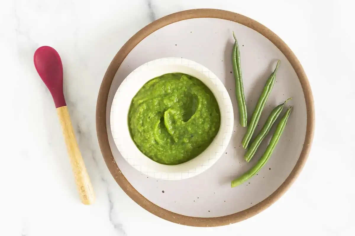 Gr&uuml;ner Brei aus Bohnen, ideal ab wann Bohnen Baby essen darf. Daneben liegen frische gr&uuml;ne Bohnen und ein L&ouml;ffel.