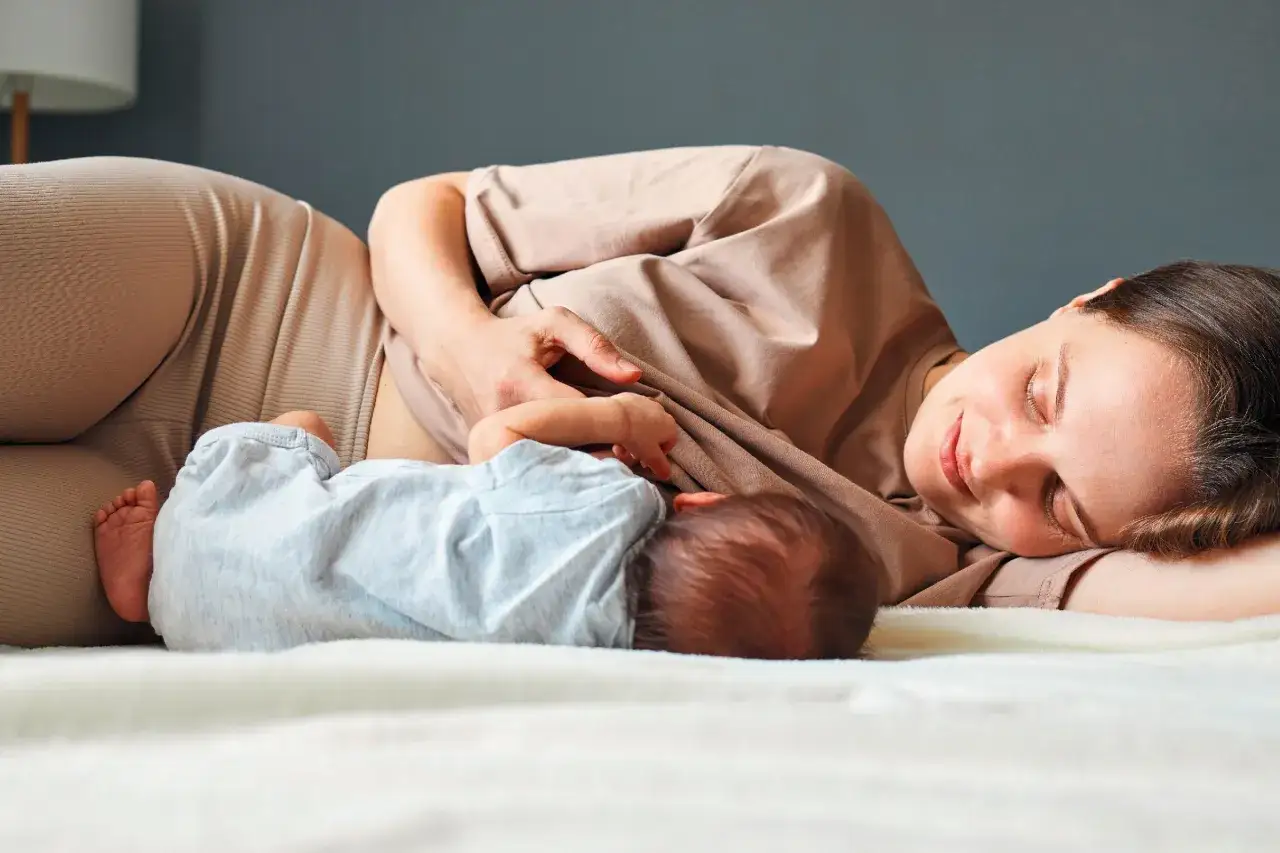 Eine Mutter liegt entspannt und lächelt, während sie ihr Baby stillt. Dies ist ein Moment des cluster feeding, der Nähe und des Wohlbefindens.