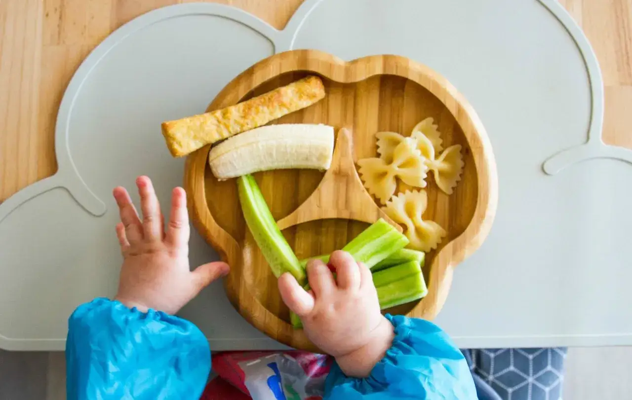 Baby is reaching for food on a plate: banana, cucumber sticks, pasta. Ab wann darf ein Baby Obst essen?