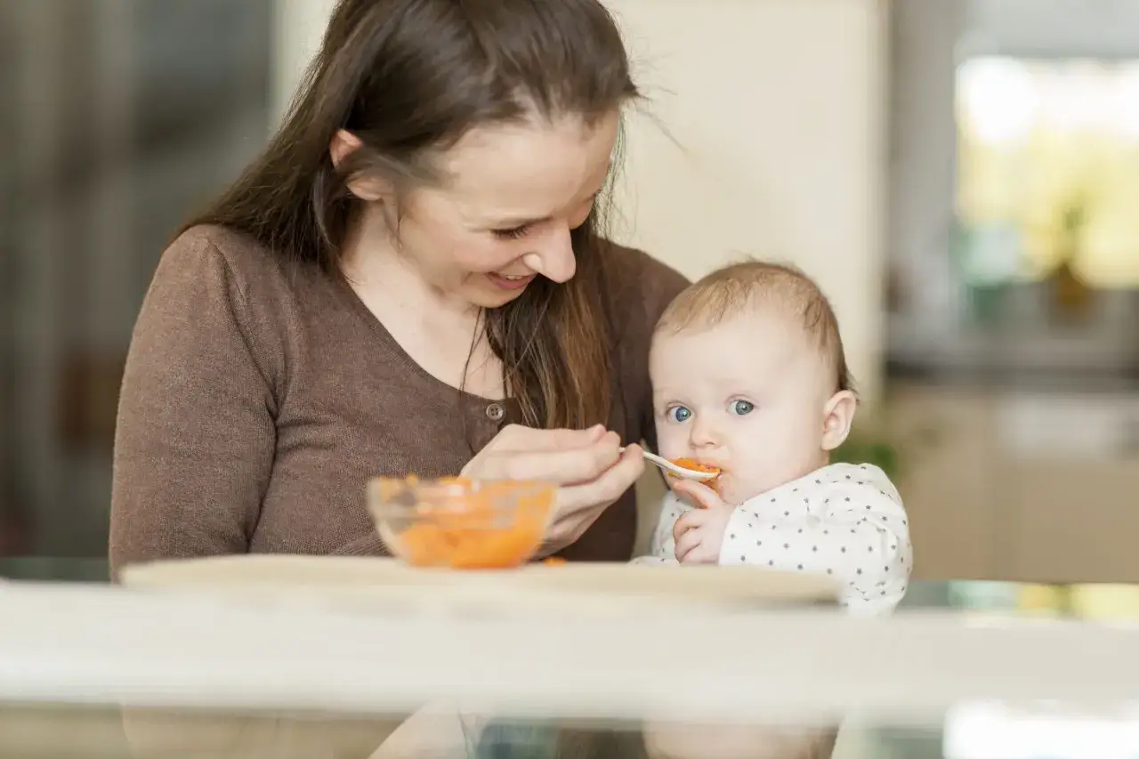 Mutter f&uuml;ttert Baby mit Karottenbrei. Ein s&uuml;&szlig;er Moment vor oder nach dem Stillen, der die ersten L&ouml;ffel fester Nahrung feiert.