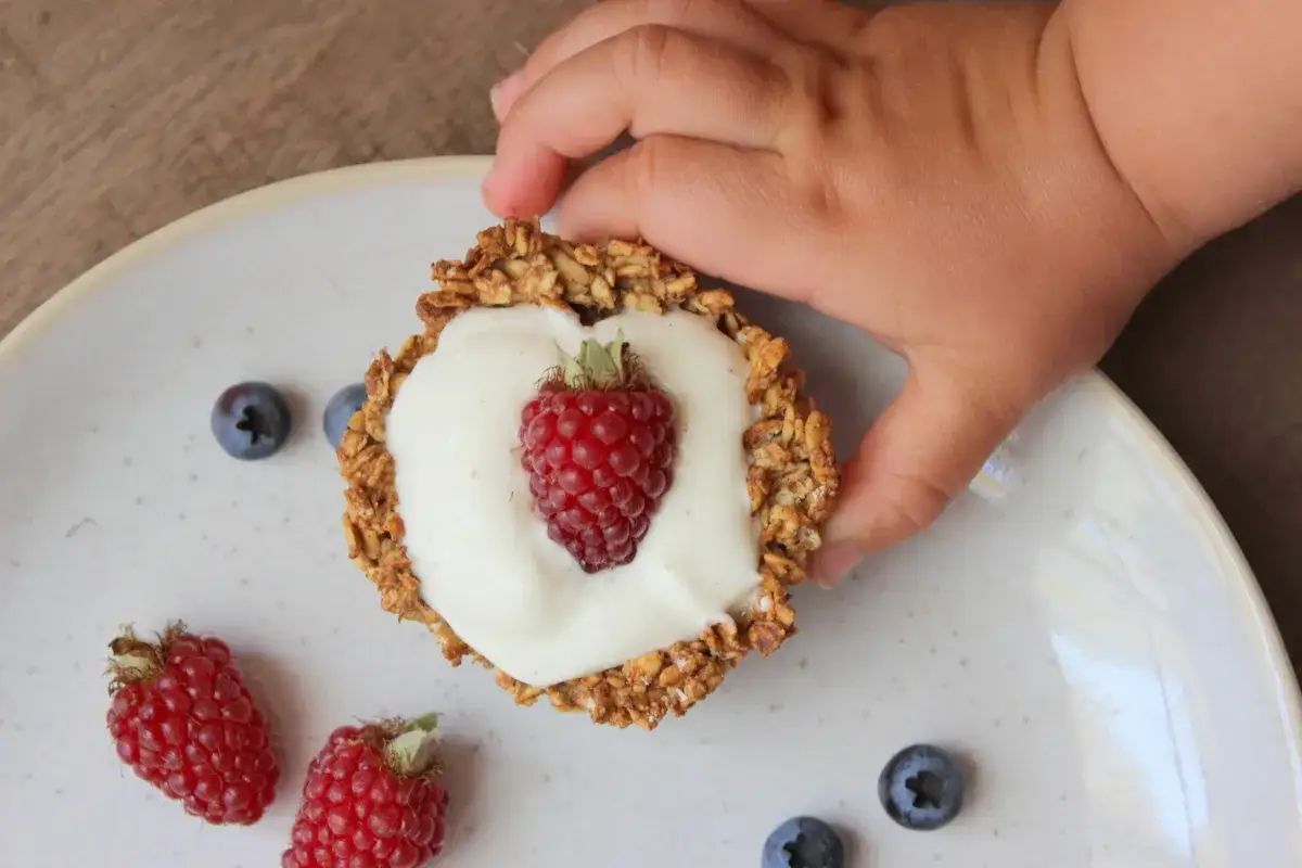 Kleinkind greift nach einem leckeren Joghurt-Granola-T&ouml;rtchen mit Himbeere und Blaubeeren.