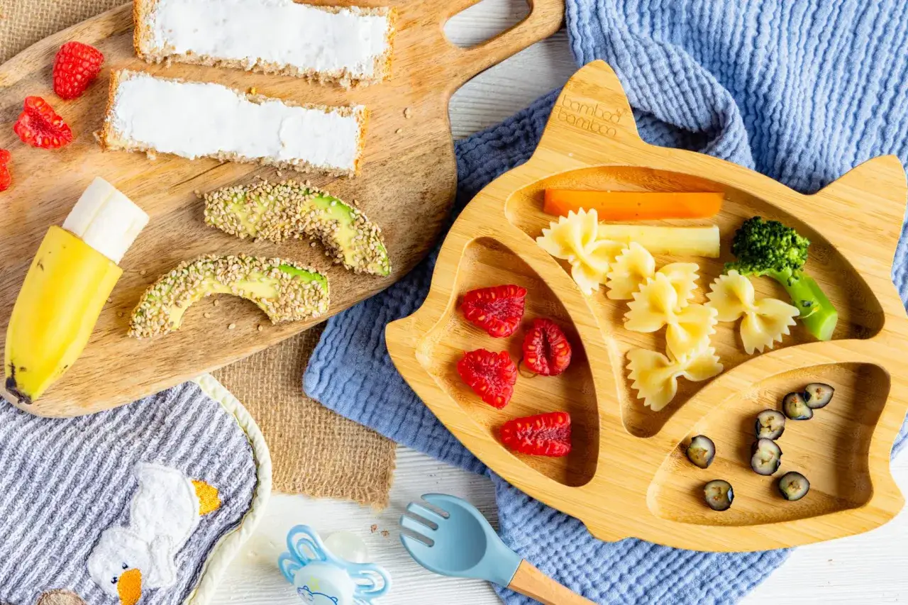 Ein blw baby entdeckt leckere Snacks: Avocado-Spalten mit Sesam, Banane, Himbeeren, Nudeln, Karotten, Brokkoli und Blaubeeren auf einem Katzen-Teller.
