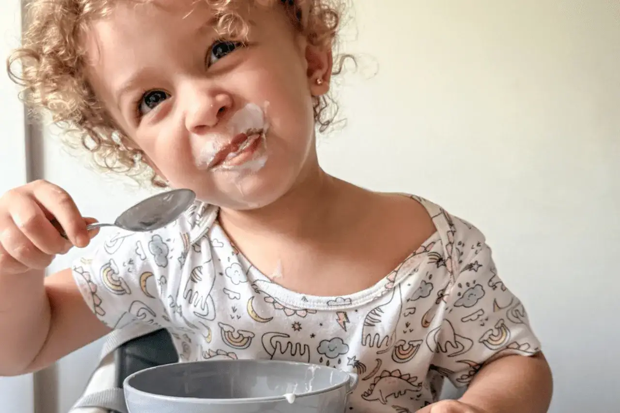 Kleines Kind mit Joghurt im Gesicht und Löffel in der Hand, genießt seinen Baby Naturjoghurt.