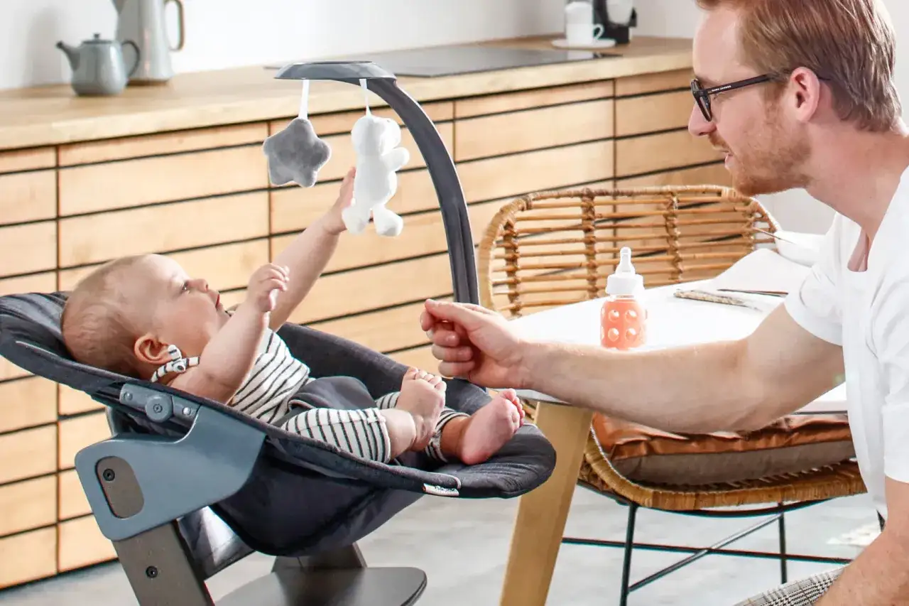 Ein Vater spielt mit seinem Baby in einer Wippe. Das Baby greift nach Spielzeug. Bald kann das Baby sitzen.