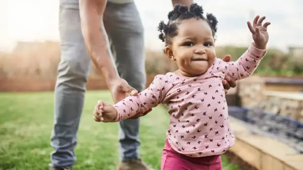 Ein Baby lernt laufen! Ein kleines Mädchen in einem rosa Shirt mit Schmetterlingen macht seine ersten Schritte im Garten, gehalten von einer erwachsenen Hand.