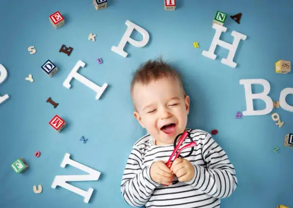 Lachendes Baby mit Brille inmitten von Buchstaben. Bald beginnt die spannende Reise, ab wann sprechen Kinder?