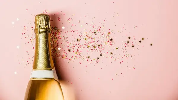 Goldene Sektflasche mit bunten Streuseln und Sternen auf rosa Hintergrund. Ein Glas Sekt stillen für den besonderen Moment.
