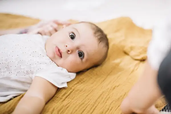 Ein neugieriges Baby, etwa 3 Monate alt, liegt auf einer senfgelben Decke und blickt mit großen Augen in die Kamera.