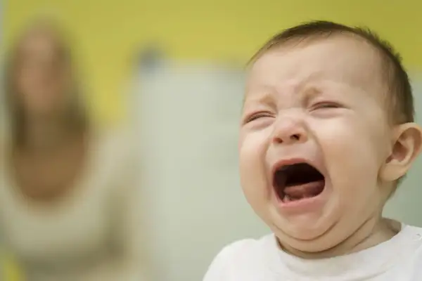 Ein Baby kreischt schrill, die Augen geschlossen, der Mund weit geöffnet. Im Hintergrund eine unscharfe Person.