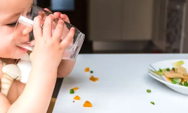 Kleines Kind trinkt aus Glas, daneben ein Teller mit Brei und verstreuten Gemüsestückchen. Wie viel Wasser bei Beikost?