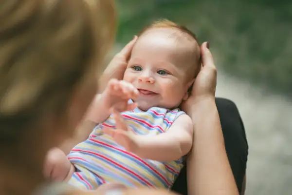 Ein lächelndes neugeborenes Baby mit blauen Augen wird von Händen gehalten. Es trägt ein gestreiftes Oberteil und streckt seine Arme aus.