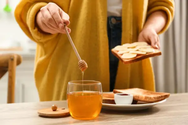 Goldener Honig tropft von einem Holzstäbchen auf ein Glas. Eine Person bereitet Toast mit Bananen und Honig zu, ein süßer Genuss beim Stillen.